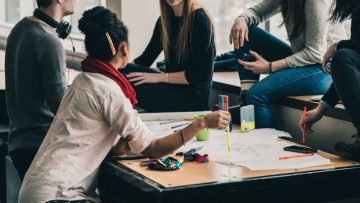 A group of young people sitting around a desk which has stationery and papers spread across it