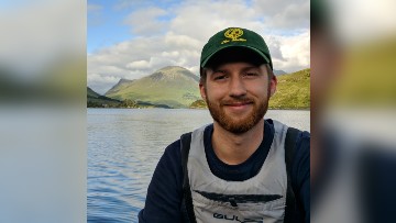 Atticus, wearing a navy sweater, grey lifejacket and green baseball cap, smiles towards the camera. A lake, mountains and blue sky are visible in the background