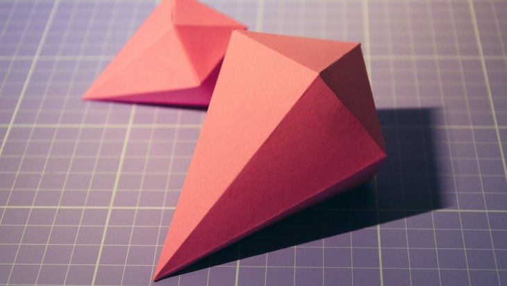 Two pieces of origami made with red-orange paper. They are laying on a gridded blue cutting mat.