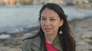 Nelly, in a red top and khaki jacket, smiles towards the camera. A beach is visible in the background.