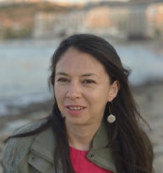 Nelly, a woman with olive skin and long, dark hair looks toward the camera. A beach and water are visible in the background.