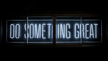 A neon sign against a dark background. The sign reads "do something great" in capital letters, and is surrounded by a plain border
