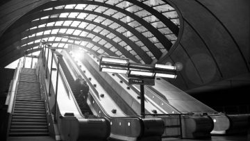 Two people ascending an escalator in a tube station. There is a large glass roof overhead and the sun is shining through. The image is in black and white.