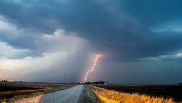 A dark cloudy sky with a lightning flash striking a country road. Yellow grass and brown earth are visible on both sides of the road.