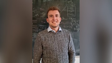 Sebastian Gilbert, a white man with blonde hair wearing a grey jumper, stands in front of a blackboard