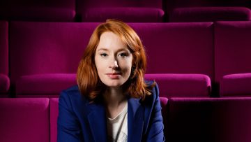 Image of Hannah Fry sitting on a row of purple auditorium seats, arms folded over her knees.