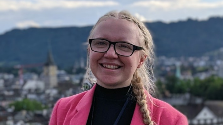 Sophie Hawkes is smiling and facing forward, wearing a pink jacket with long plaited fair hair and glasses